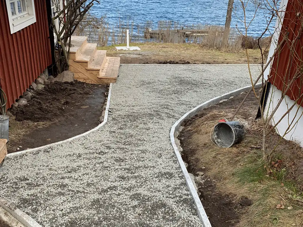 Bilden visar en nyanlagd grusgång som leder fram till en byggnad, med en tydlig, rundad kant som ger en mjuk och naturlig känsla. Trappan i trä på vänster sida är en elegant detalj som kompletterar den omgivande landskapsdesignen, som är nära vattnet och ger en lugn och harmonisk atmosfär. Grusvägen och trappan skapar en funktionell och estetiskt tilltalande ingång, perfekt för både privata och offentliga fastigheter. Denna typ av stenläggning ger en naturlig känsla och passar väl in i utomhusmiljöer med mycket grönska och vattennära områden. Bilden visar en nyanlagd grusgång som leder fram till en byggnad, med en tydlig, rundad kant som ger en mjuk och naturlig känsla. Trappan i trä på vänster sida är en elegant detalj som kompletterar den omgivande landskapsdesignen, som är nära vattnet och ger en lugn och harmonisk atmosfär. Grusvägen och trappan skapar en funktionell och estetiskt tilltalande ingång, perfekt för både privata och offentliga fastigheter. Denna typ av stenläggning ger en naturlig känsla och passar väl in i utomhusmiljöer med mycket grönska och vattennära områden.
