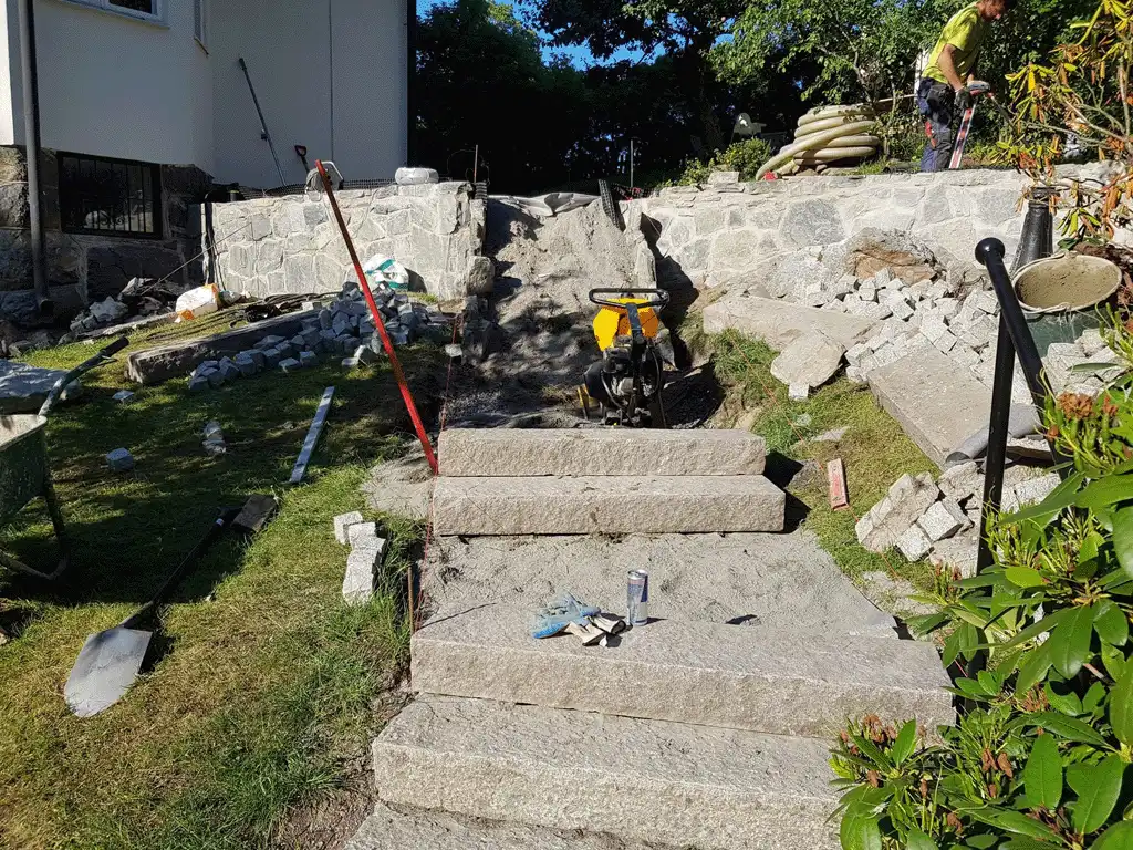 Bilden visar en byggarbetsplats där en stentrappa byggs. Trappan är gjord av stora stenplattor, och det omgivande området är under landskapsarbete. Flera byggmaterial, inklusive stenfragment och verktyg, ligger utspridda runt platsen. I bakgrunden syns arbetare som slutför projektet, med vissa delar fortfarande under konstruktion. Stenläggningen är stadig och välformad, och den övergripande designen tar form, vilket lovar ett vackert resultat när det är klart. Bilden visar en byggarbetsplats där en stentrappa byggs. Trappan är gjord av stora stenplattor, och det omgivande området är under landskapsarbete. Flera byggmaterial, inklusive stenfragment och verktyg, ligger utspridda runt platsen. I bakgrunden syns arbetare som slutför projektet, med vissa delar fortfarande under konstruktion. Stenläggningen är stadig och välformad, och den övergripande designen tar form, vilket lovar ett vackert resultat när det är klart.